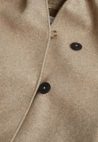 Cappotto di lana beige con due bottoni neri visibili e un fermaglio ad anello su un tessuto morbido e testurizzato.