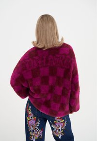 Měkký, velký kostkovaný svetr v odstínech fuchsiové a vínové barvy s jemným logem. Kombinován s tmavými džínami s barevnými vyšívanými vzory.