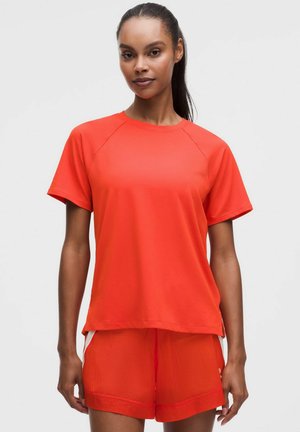 Vrouw met donker haar in een paardenstaart, die een losse feloranje korte mouwen T-shirt en bijpassende korte broek draagt, staand tegen een effen achtergrond.