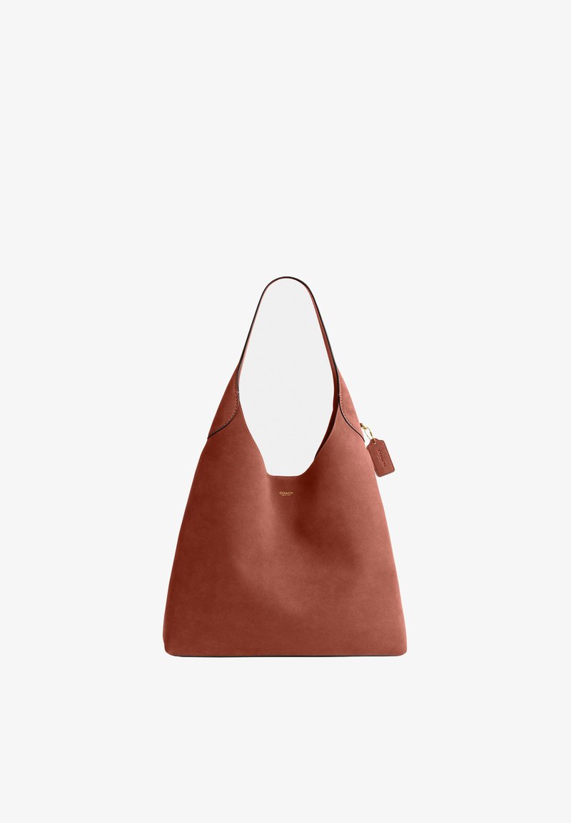 Sac cabas en cuir marron avec une large bandoulière unique, petit logo Coach doré et étiquette en cuir attachée sur fond blanc.