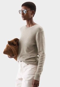 Suéter gris claro con cuello redondo y puños acanalados, combinado con una chaqueta de ante marrón. El modelo lleva gafas de sol de gran tamaño. Pantalones blancos.