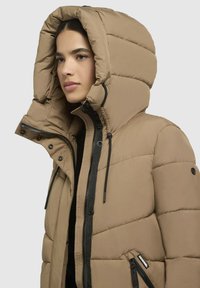 Giacca a vento color beige, dotata di un ampio cappuccio, tasche con zip e un design testurizzato trapuntato con accenti neri e cordini.