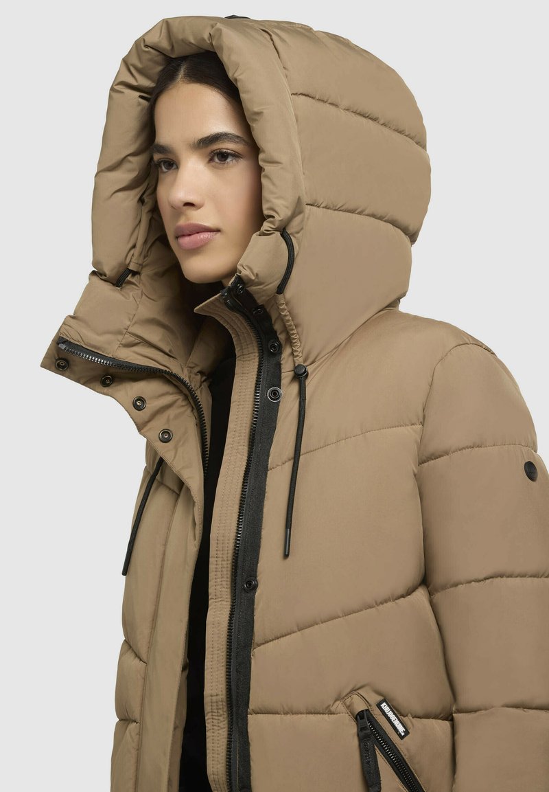 Tan pufferjack met een grote capuchon, ritszakken en een gestructureerd, gewatteerd ontwerp met zwarte accenten en trekkoorden.