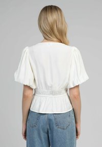 Femme aux cheveux blonds portant un blouse blanche à manches bouffantes avec taille élastique, associée à un jean en denim bleu taille haute, vue de dos.