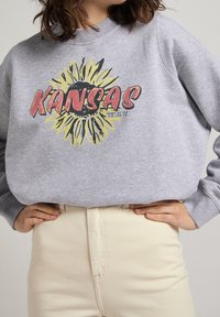 Felpa grigia con un grande girasole e il testo "KANSAS", indossata da una persona con le mani sui fianchi, abbinata a pantaloni beige chiaro.