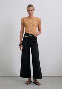 Texturiertes, orangefarbenes Crop-Top mit kurzen Ärmeln, kombiniert mit hoch taillierten, weit geschnittenen schwarzen Jeans mit dekorativen Knopfdetails. Schwarze Flip-Flops.
