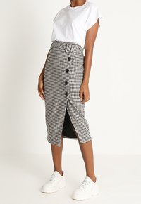 Falda midi de cuadros en blanco y negro con cierre de botones en la parte delantera, cintura alta y abertura lateral. Combinada con una camiseta blanca de manga corta y zapatillas.