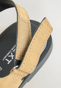 Lanière de sandale en daim beige avec fermeture Velcro sur une semelle noire, affichant un texte imprimé partiel.