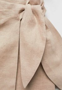 Beige linnen stof met een knoopdetail, met een brede, vloeiende strik en een zachte textuur, wat een gedrapeerd uiterlijk creëert.