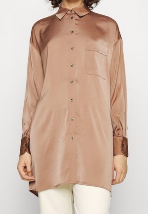 Kobieta ubrana w luźną, satynową koszulę na guziki z długim rękawem w kolorze przydymionego różu z kieszenią na piersi, w połączeniu z jasnymi spodniami.
