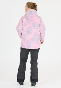 Wasserdichte pinke Jacke mit einem strukturierten, pastellfarbenen Aquarelldesign, kombiniert mit schwarzen Hosen, die an den Knöcheln mit Reißverschlüssen für Anpassungsfähigkeit versehen sind.