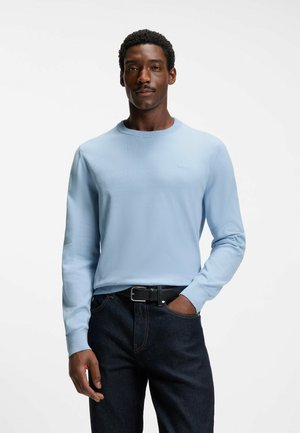 Mann trägt hellblauen Pullover mit langen Ärmeln und dunkle Jeans, steht mit einer Hand in der Tasche vor einem einfarbigen weißen Hintergrund.