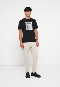 T-shirt en coton noir avec un graphique central d'un ours rugissant, associé à un pantalon beige clair et des mocassins noirs.
