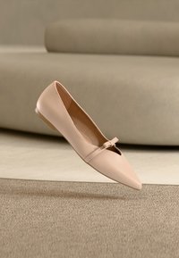Ballerine beige en cuir à bout pointu, avec une bride décorative, une texture lisse et un petit talon, suspendue au-dessus d'une surface texturée.
