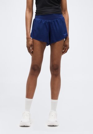 Femeie purtând pantaloni scurți sportivi bleumarin, bluză neagră, șosete albe scurte și încălțăminte sport albă, stând în picioare pe un fundal simplu.