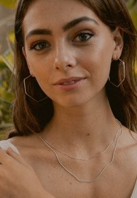 Silberne Halskette mit zwei geschwungenen Ketten in Schichten, die geometrische Creolenohrringe ergänzt. Das Modell hat Wassertropfen auf der Haut und trägt ein weißes Oberteil.