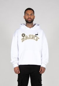 Witte hoodie met geborduurd "SAINT" logo in beige, voorzien van een voorzak en een capuchon met trekkoord, gedragen met zwarte cargobroek.