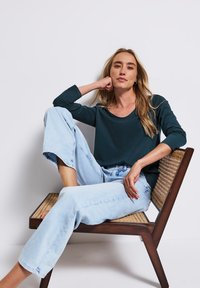 Donkergroene langemouwtop gecombineerd met lichtblauwe brede jeans, zittend op een geweven stoel, met lang blond haar en een ontspannen houding.