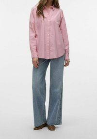 Camisa rosa de botón con cuello clásico, mangas largas y un corte relajado, combinada con vaqueros de pierna ancha de color azul claro y zapatos marrones.