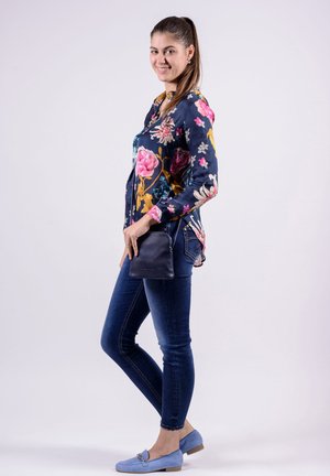 Junge Frau lächelnd, seitlich stehend, trägt eine geblümte Bluse, dunkle Skinny-Jeans, blaue Loafers und hält eine kleine marineblaue Handtasche.