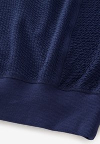 Tissu bleu foncé avec un motif tricoté texturé et ondulé et un ourlet côtelé en bas, suggérant un détail de vêtement.