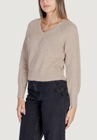 Pullover beige a maglia con scollo a V, dotato di texture a pois e bordo a costine. Maniche lunghe con polsini aderenti, abbinato a pantaloni neri scuri.