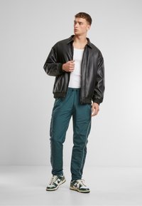 Chaqueta bomber de cuero negra, camiseta sin mangas blanca, pantalones de chándal turquesa con franjas laterales, y zapatillas verdes y blancas. Fondo liso.