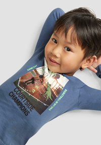 Langärmliges blaues T-Shirt mit einem Grafikdruck, der eine Dinosaurier- und Basketballszene zeigt. Der Text lautet "Courtside Champions" und "Game Day."