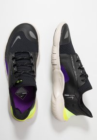 Un paio di scarpe da corsa Nike nere, viola e giallo neon con suole beige, lacci visibili e testo che indica l'impermeabilità e la riflettività.