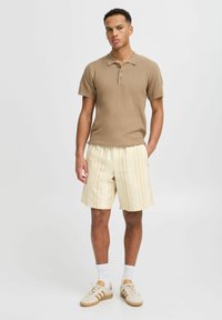 Camisa de polo corta de mangas en color marrón con tejido texturizado, combinada con pantalones cortos a rayas amarillas claras. El modelo lleva calcetines blancos y zapatillas ligeras.