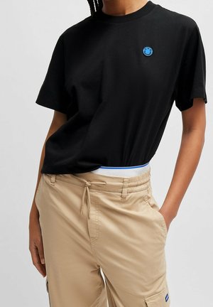 Henkilö, jolla on musta t-paita, jossa pieni sininen hymyilevä logo, sekä beiget nauhakuoriset housut sivutaskuilla, käsi taskussa, studiotausta.