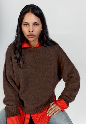 Femme aux longs cheveux foncés portant un pull marron par-dessus une chemise rouge rayée et un jean bleu clair, assise avec une main sur la hanche.