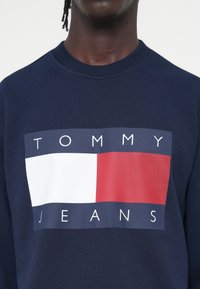 Felpa blu navy con una grande grafica che riporta il testo "TOMMY" sopra un rettangolo bianco e rosso, e "JEANS" sotto. Realizzata in cotone e con collo a girocollo.
