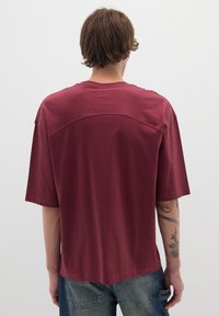 T-shirt oversize bordeaux con maniche corte e scollo rotondo. Materiale in cotone morbido, caratterizzato da un sottile dettaglio di cucitura sulla parte superiore della schiena.