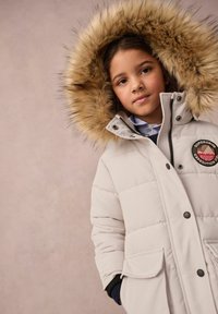 Hellgraue gesteppte Jacke mit einer abnehmbaren Kunstfellkapuze, Fronttaschen und einem Logo-Patch. Die Jacke hat einen Reißverschluss und einen Knopfverschluss.