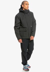 Schwarze wasserdichte Jacke mit Kapuze, mit Reißverschluss vorne, zwei Taschen vorne und verstellbaren Bündchen. Getragen mit dunklen Hosen und bunten Turnschuhen.