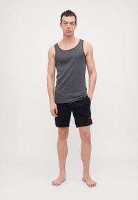 Tricou gri fără mâneci din bumbac, asociat cu pantaloni scurți negri care au un patch cu un logo roșu. Modelul stă desculț pe un fundal simplu.