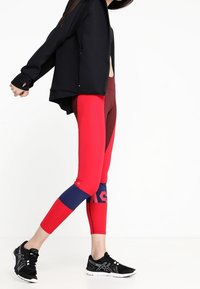 Czerwone i bordowe legginsy z teksturowanym wzorem, z granatowymi akcentami i dopasowanym krojem. W zestawie z czarną kurtką zapinaną z przodu i czarnymi sneakersami.