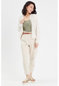 Cardigan en dentelle léger de couleur crème, porté sur un débardeur ajusté vert olive. Associé à un pantalon beige fuselé et des sandales dorées métalliques à brides.