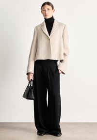 Μπεζ cropped σακάκι με υφή, κλείνει με ένα κουμπί, συνδυασμένο με μαύρο παντελόνι φαρδιάς γραμμής και μικρή μαύρη τσάντα χειρός.
