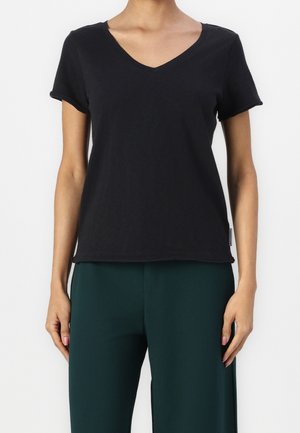 Mujer con camiseta negra ajustada de manga corta y escote en V, y pantalones de cintura alta de color verde oscuro, de pie contra un fondo blanco.