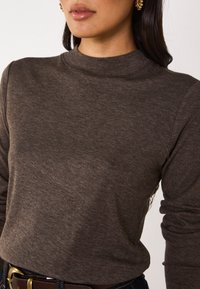 Maglietta marrone a maniche lunghe con un colletto alto e un tessuto morbido e strutturato. Indossata con una cintura scura, esaltando una silhouette aderente.