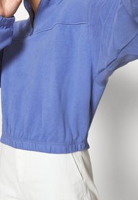 Sudadera corta de color azul claro con un cuello de media cremallera, cinturilla elástica y mangas abullonadas. La tela parece suave con una textura lisa.