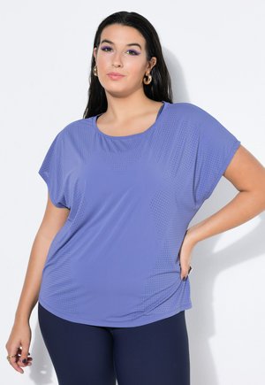 Pitkätuukkainen nainen, jolla on löysä laventelinvärinen rei'itetty lyhythihainen paita ja tummat trikoot, seisoo yhdellä kädellä lanteilla.