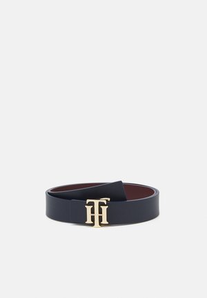 Ceinture en cuir noir avec une boucle de ceinture dorée entrelacée formant le logo "TH", posée à plat sur un fond blanc.