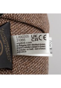 Clothing label on brown textured fabric with product code, website, UKCA and CE marks, and origin information showing eco-friendly production.