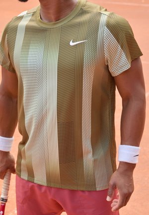 Muž nosící pruhované olivově zelené a béžové tričko Nike a růžové kraťasy držící tenisovou raketu na antukovém kurtu.