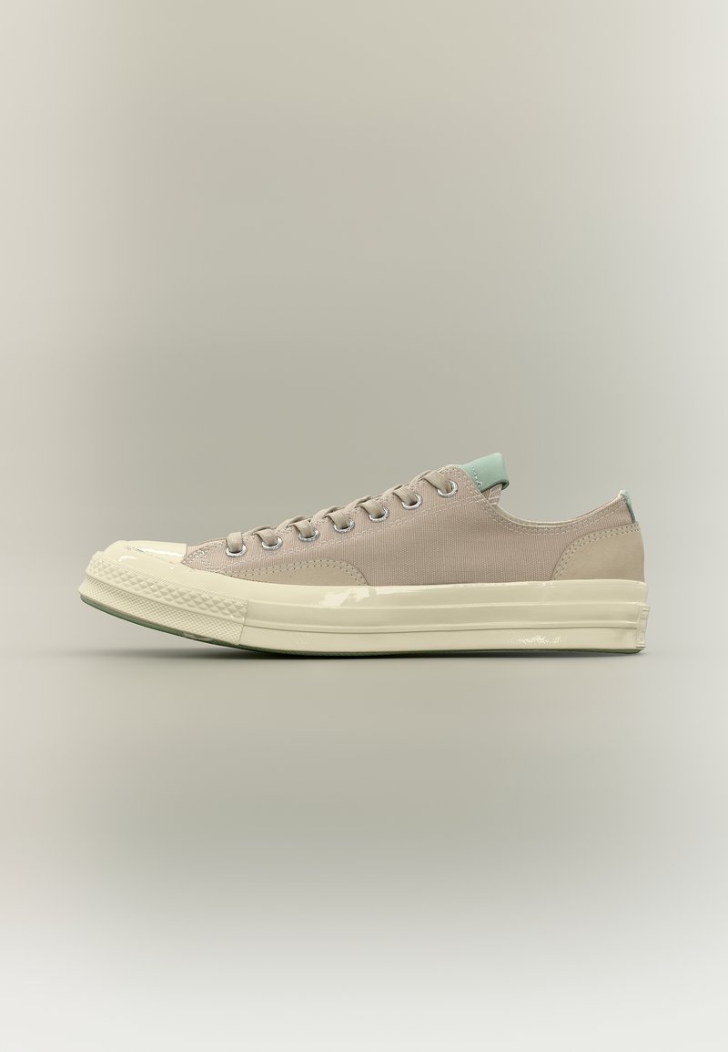 Μπεζ χαμηλό sneaker με κρέμα σόλα από καουτσούκ και προστατευτικό άκρου, ανοιχτοπράσινη ετικέτα στη φτέρνα και στη γλώσσα, ορατές μεταλλικές τρύπες για τα κορδόνια και υφασμάτινο επάνω μέρος.