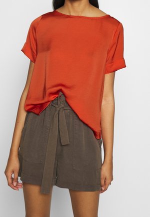 Roest-oranje blouse met korte mouwen en een ronde halslijn, gecombineerd met hoge kaki shorts met een gestrikte taille en zijzakken.