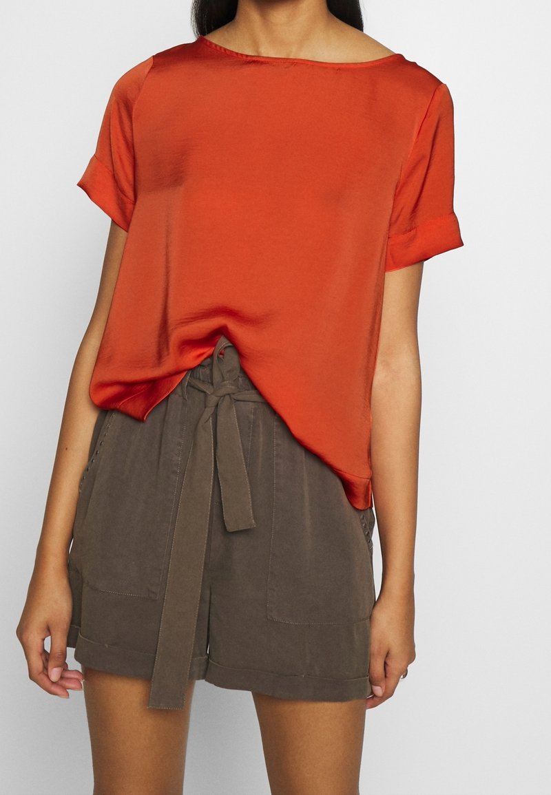Blusa corta a maniche corte di colore ruggine, con scollatura rotonda, abbinata a pantaloni corti khaki a vita alta con cintura annodata e tasche laterali.